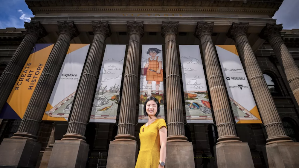 Artist Natasha Sim standing in front of the State Library of Victoria with her #CreateArtHistory banners displayed between the columns